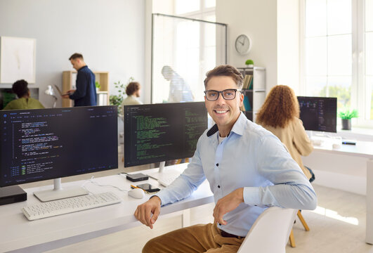 Developer programmer man smiling in office workstation. He works at two monitors with coding on computer, working with a team in a bright open space. Concept of teamwork, innovation, and success.