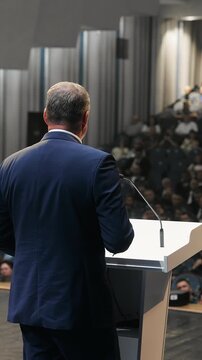 Male business speaker gives a presentation at a podium in front of a large audience during a professional conference or corporate event.