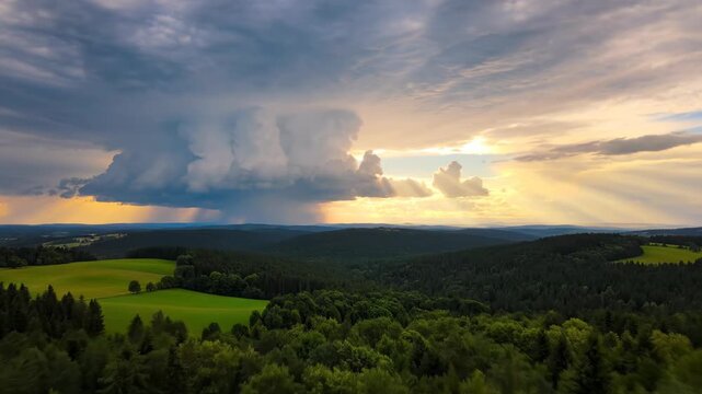 Aerial view of rolling green hills and dense forests under a dramatic sunset sky with a massive cumulonimbus cloud, heavy rain shaft, and bright golden sunbeams across the horizon.