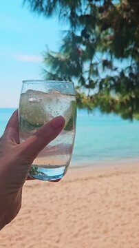 Female hand holding cold beverage or cocktail by the beach during holiday. Enjoying refreshing drink while on vacation in greek island. POV from phone in vertical UGC style. Leisure mode in paradise