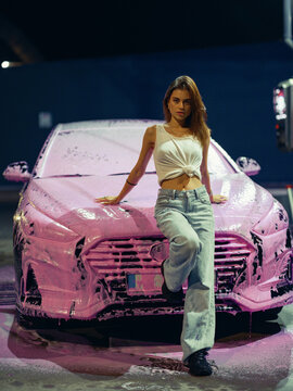 Young Woman Leaning On Hood Of Car Covered In Pink Foam At Night Self Service Wash Station