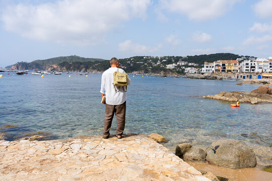 Man on a pier with a backpack and a book, looking at the clear water and coastal village of Calella de Palafrugell