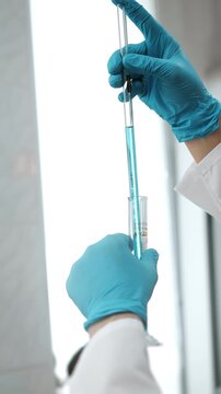 Close-up of laboratory technician measuring blue liquid with a graduated glass pipette during precise scientific analysis and sample preparation.