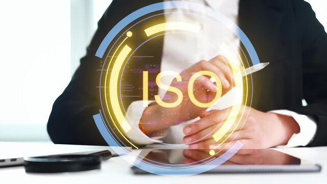 A person is sitting at a desk in a bright office. They hold a pen while discussing ISO standards. A tablet is on the desk and a circular graphic shows the ISO logo.