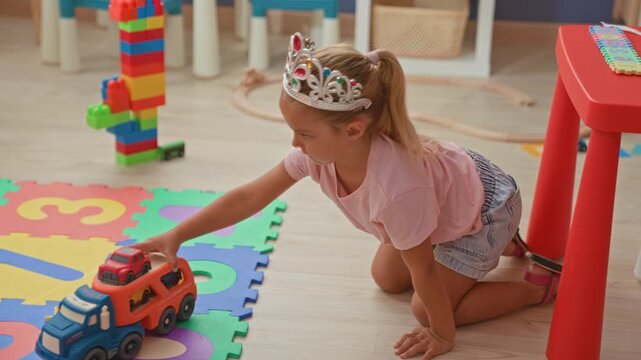 Girl reaches hand for toy transport truck on colorful foam playmat among blocks and bucket inside a building playroom, kneeling and leaning forward; playful.
