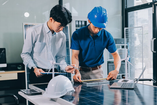 Two Asian engineers wearing hardhats and safety vests discussing solar panel installation and renewable energy project in the office.