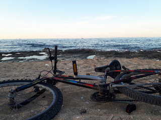 Mountain bike lying on its side on sandy seashore beside rocky coastline at dusk. Calm sea and pastel sky evoke adventure, freedom, and outdoor travel themes for cycling campaigns. © Grenar