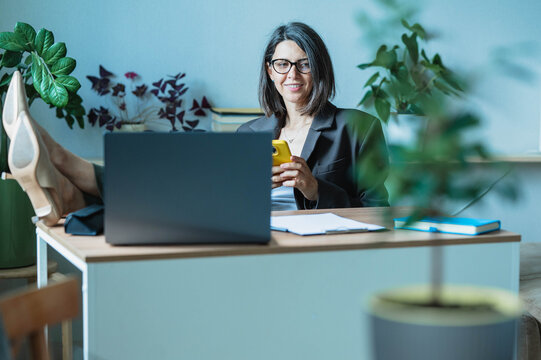 Businesswoman relaxes with feet on desk and uses smartphone at home