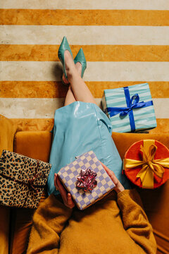 A woman sits on a couch indoors unwrapping a Christmas gift