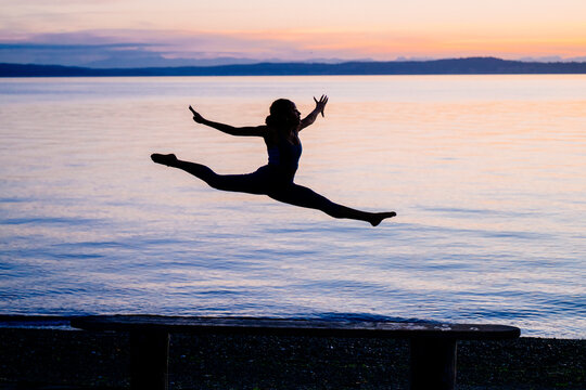 Hispanic woman performs split jump silhouette at sunset over lake