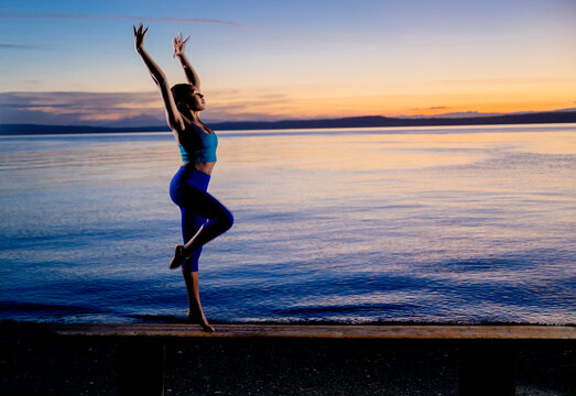 Hispanic woman practicing yoga balance pose on bench by lake at sunset