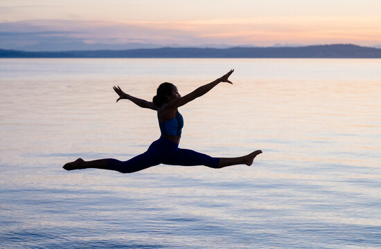 Hispanic woman performing jumping split yoga pose over lake at sunset