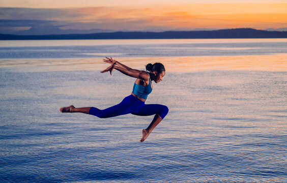 Hispanic woman performing a split jump exercise over lake at sunset