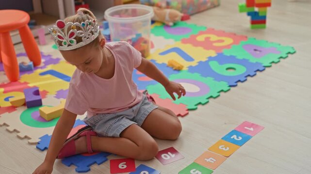 Girl arranging number cards with her hand on foam mat inside a building playroom; playful curiosity.