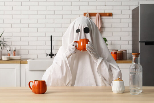 Halloween ghost costume drinking coffee in a modern kitchen, spooky morning routine and holiday celebration