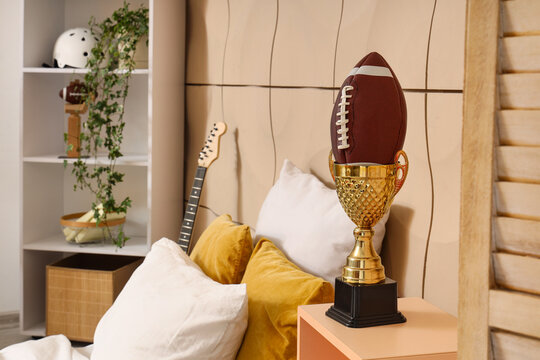 Sports achievement and victory, American football ball inside a golden trophy cup on a bedroom nightstand with hobby items in the background