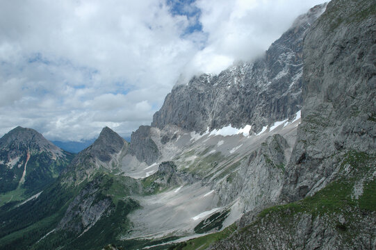 Dramatic rocky peaks and steep walls of the Dachstein mountains rising above a valley with clouds and snow patches in Austria