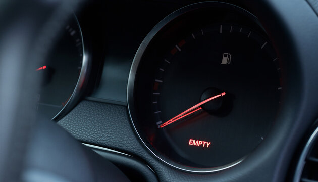 Car fuel gauge needle pointing to empty with red illuminated warning light glowing on dark black automotive dashboard closeup