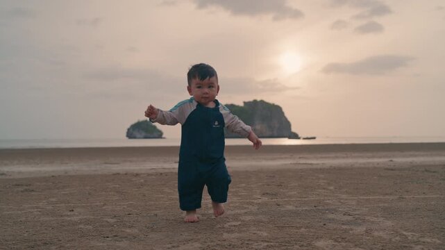 Cute Asian toddler taking steps barefoot towards camera on sandy beach at golden hour.