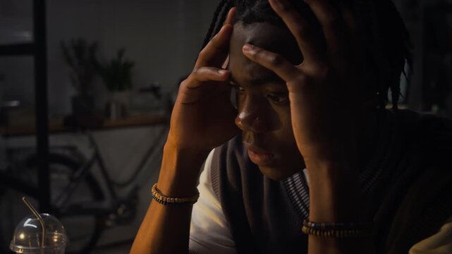 Close up of African American young adult man holding head in hands while working late at computer screen in dark office. Video suitable for content on workplace stress, burnout and overtime
