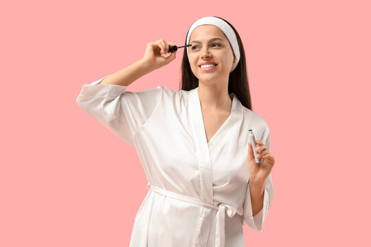 Pretty young woman in bathrobe and headband applying mascara on pink background