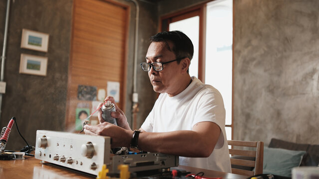 Mature Asian man disassembling and checking audio equipment, spraying contact cleaner, cleaning circuit board for repair, home appliance DIY fix, electronics component hobby, handyman skill workshop.