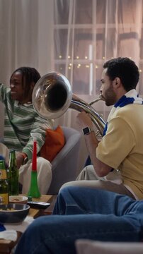 Vertical video of multiethnic young adult male football fans cheering and playing brass instruments at home while watching sports. Perfect for sports fandom and streaming promotions