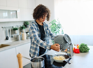 A young person teenager boy cooks and drains pasta in a bright, modern kitchen, steam rising as...