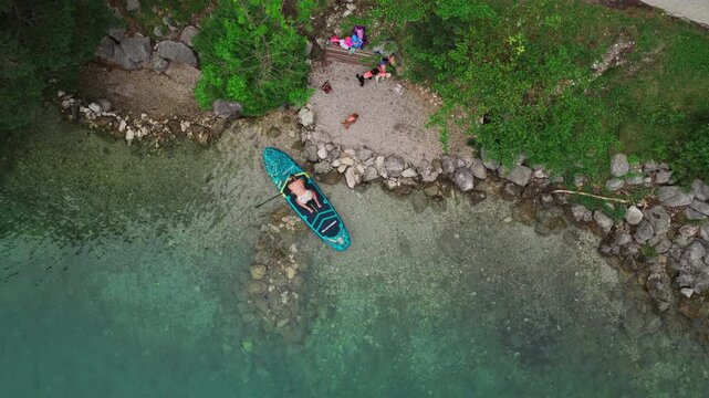 An inflatable stand up paddle board floats beside the rocky shoreline of Achen Lake in Tyrol, Austria, with clear lake water and a rugged natural shore visible - drone top down