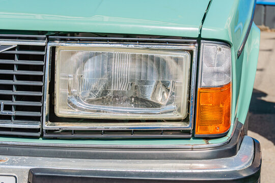 Vintage classic car front section showcasing headlight, grille, and body paint in light teal, featuring worn chrome trim, rusted slats, and faded color against an out-of-focus outdoor setting on a