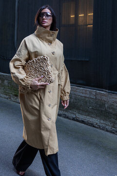 Elegant urban fashion portrait featuring confident woman in minimalist beige outerwear, wide trousers and statement chain bag against dark architectural background