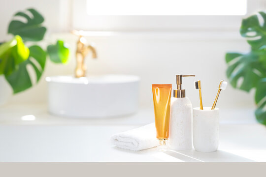White table top with soap dispenser, amber color toothpaste tube, toothbrush holder and folded spa towel on bright blurred bathroom with white vessel sink and gold faucet background