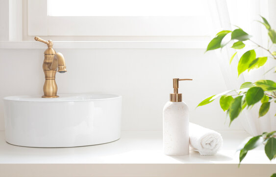 Bright white bathroom sink with bronze faucet, soap dispenser, folded spa towel and green foliage on blurred curtained window sill background