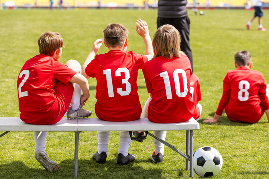 Youth Soccer Players in Red Numbered Kits Sitting on Sideline Bench and Grass Watching Game - Junior Football Team Management