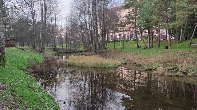 A small river flows among grassy banks, on which trees grow and are reflected in the water. There is a benches and gazebo on the shore. There are buildings nearby. There are algae in the clean water