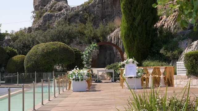 A garden is prepared for a wedding ceremony. Tables are set with flowers. The area by the pool is decorated. Rocks and trees surround the space.