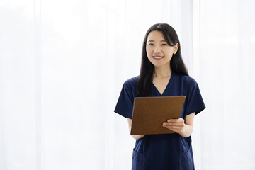 Obraz premium Young Japanese female doctor in scrubs holding a clipboard and smiling at the camera by a bright window