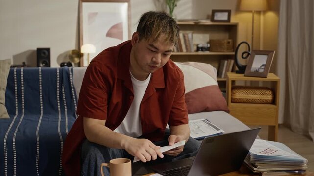 Asian young adult man sitting on floor, opening envelope and reviewing utility bills with laptop on table. Footage is ideal for commercial videos about personal finance or home budgeting
