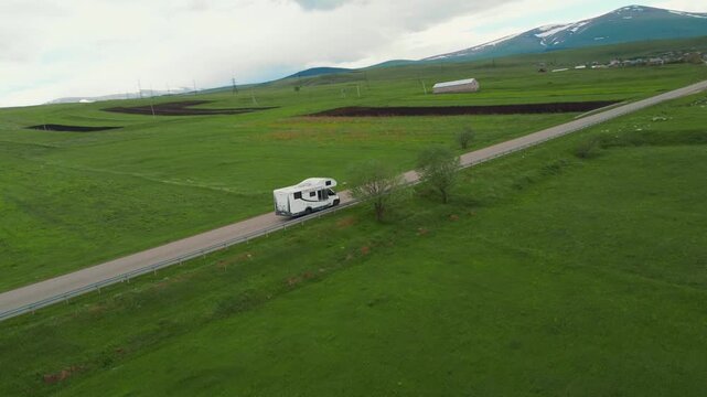 A white camper rides on an empty asphalt road through a vast green landscape with snow-capped mountains in the background, embodying the concept of freedom, travel and adventure vacation in Georgia