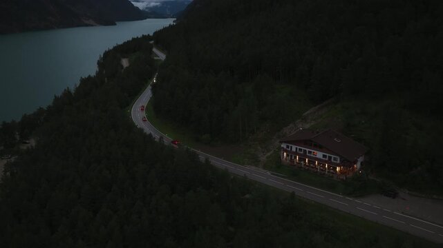 A winding road follows the shoreline of Achen Lake in Tyrol, Austria, viewed from above by a drone with the lake and surrounding landscape visible at dusk