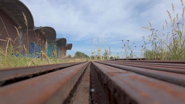 Slow dolly shot along rusty industrial tracks towards old port cranes in an abandoned harbor area