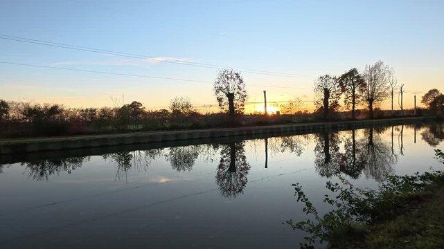 Serene canal at sunset with silhouetted trees perfectly reflected in the calm water during golden hour.
