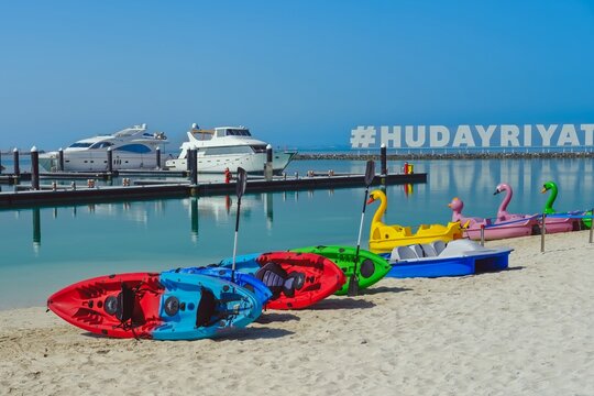 ABU DHABI, UAE, 04.03, 2021: Colorful kayaks and pedal boats line a sandy beach beside yachts and calm marina waters at Hudayriyat Island.