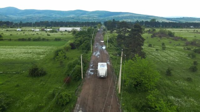 Aerial footage follows a white motorhome driving down a bumpy, wet dirt road through vast green terrain with hills in the background