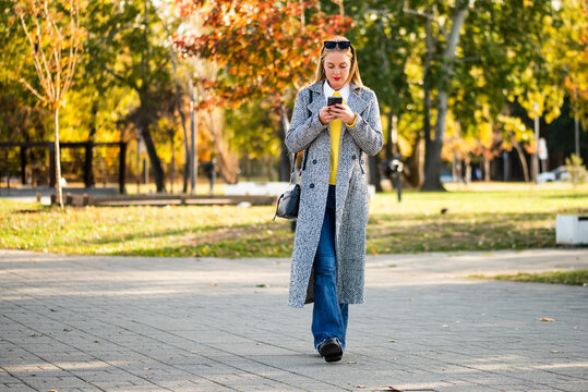 Professional woman in fashionable coat walking through autumn park and using mobile phone, representing connection, technology and busy modern lifestyle.