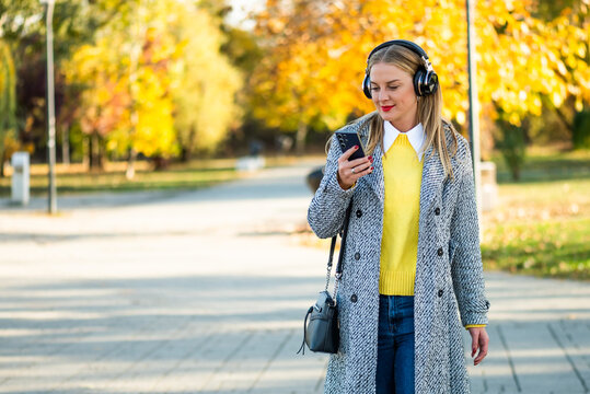 Young professional woman walking through the city park in autumn, enjoying music through wireless headphones while staying connected on her smartphone.