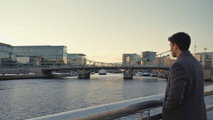 Obraz premium Man Gazing at Dublin Skyline at Dusk, Reflective Moment by the River Liffey, Contemporary Urban Scene.