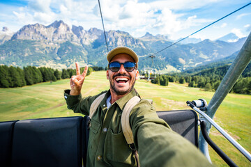 Fototapeta premium Happy young man taking selfie on chairlift during summer mountain adventure in the alps - Cheerful male smiling at camera on cable car with scenic Alpine mountains on background