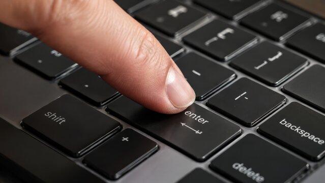 Macro close-up of a person's index finger pressing the enter key on a dark black laptop keyboard with backlit shift and delete keys visible in dramatic low-key studio lighting