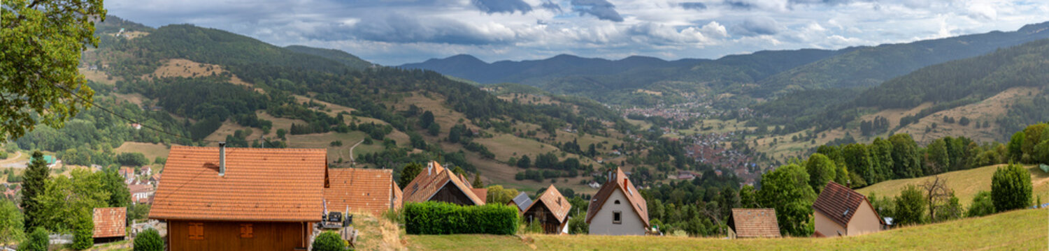 Munster, France - 08 20 2026: Panoramic view of houses on the mountain with the Munster Valley and the city in the High Vosges massif with forests and cloudy sky behind